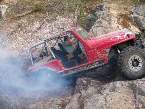 2001 Dodge 1500 - A red Jeep Wrangler with flame decals and a roll cage is shown off-roading over rocks, with smoke coming from its rear tire.