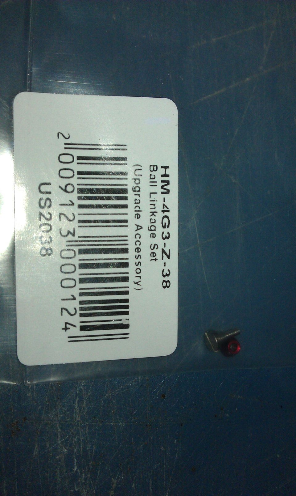 Part in plastic packaging with barcode and text 'HM-4G3-Z-38 Ball Linkage Set (Upgrade Accessory)'. Small metal part with red bead visible