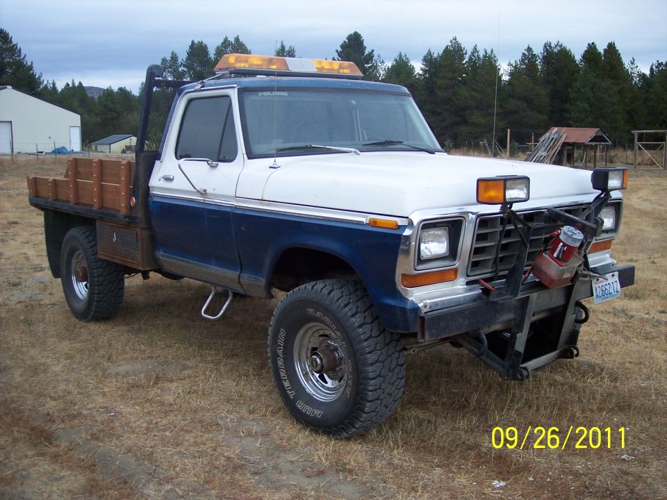 White and blue Ford flatbed pickup with roof light bar and brush guard in a field.