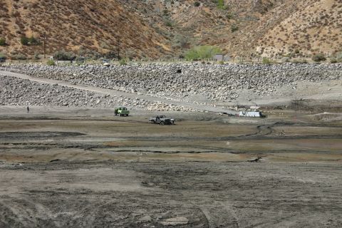 1991 Ford Bronco - Two off-road vehicles are seen in a vast, dry lake bed with a rocky embankment and sparse vegetation.