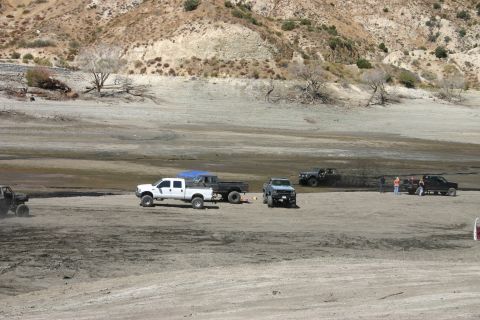 1991 Ford Bronco - A collection of trucks and off-road vehicles are seen in a wide, dry, dusty landscape with hills in the background.