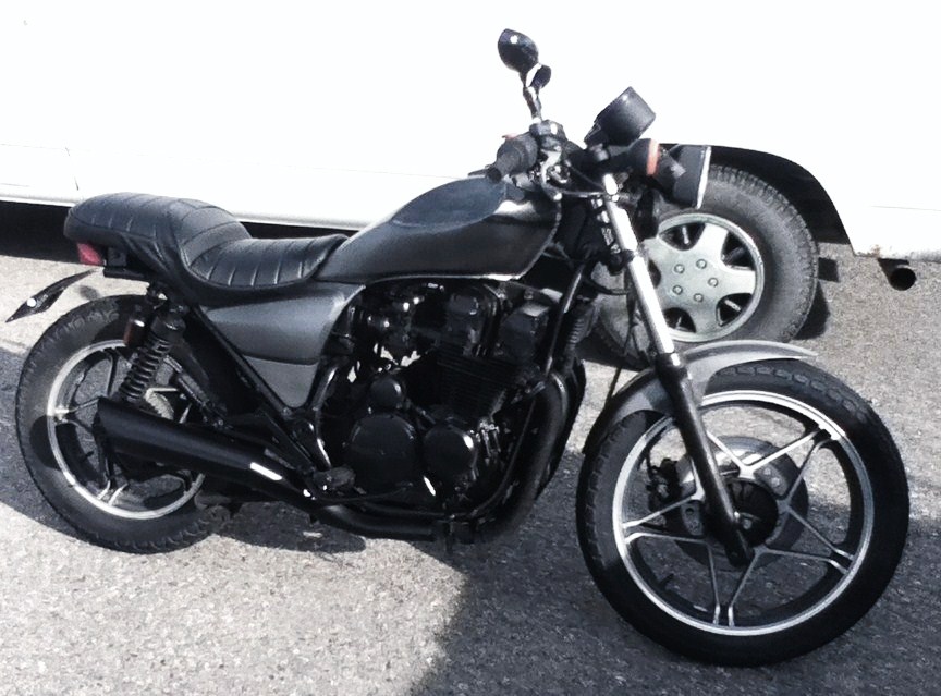 Dark colored motorcycle with a black seat and exhaust parked next to the wheel of a white vehicle