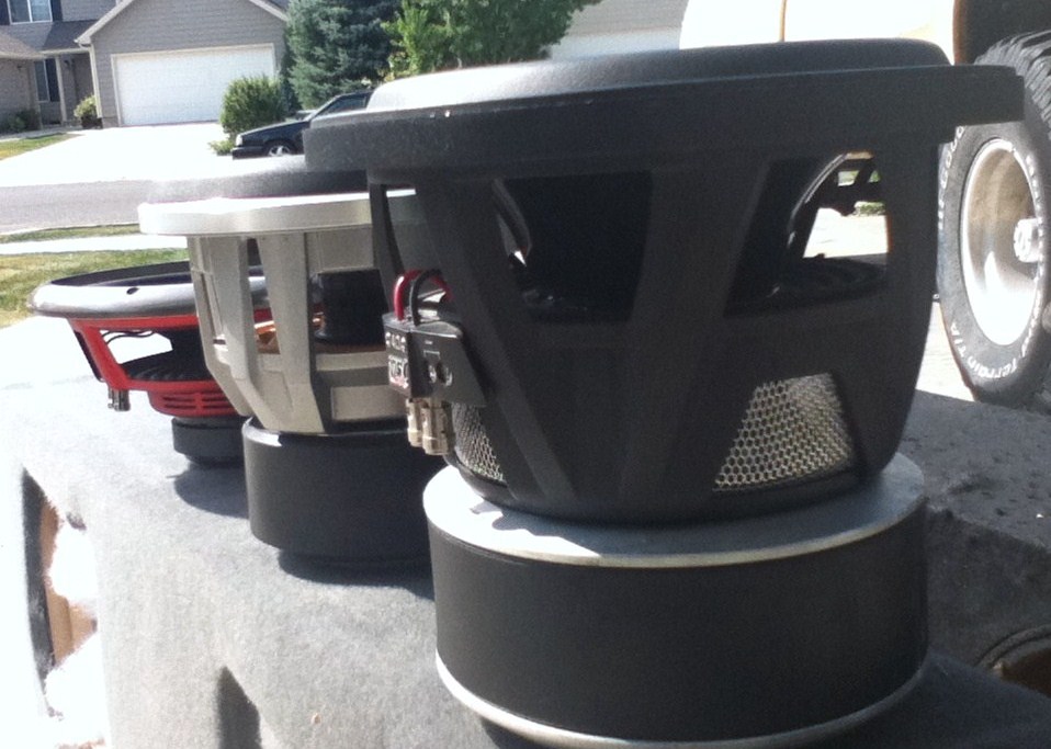 Three subwoofers lined up on a gray surface, with a house and trees in the background