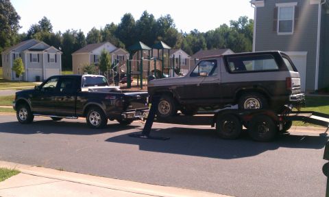 1985 Ford Bronco - A black Ford F-150 tows a trailer carrying a two-tone brown and gray Ford Bronco down a suburban street.