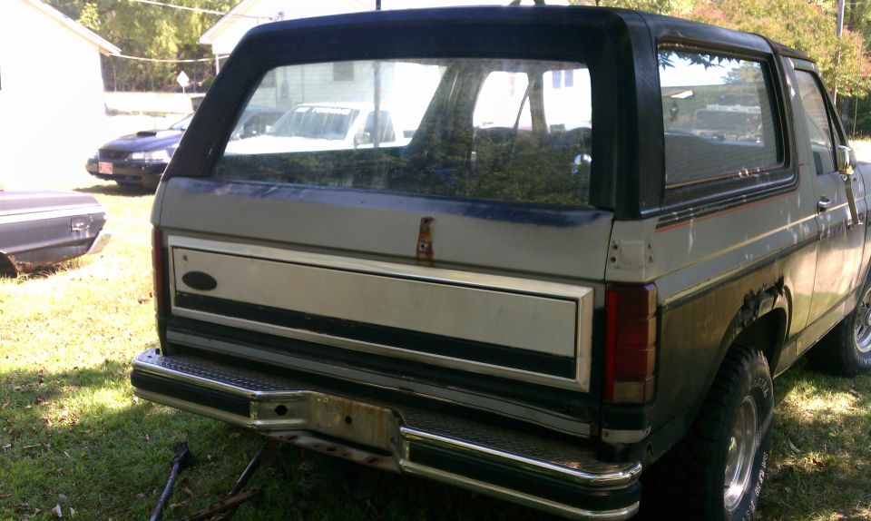 Rear view of a Ford Bronco with a black top. Another car is parked behind it