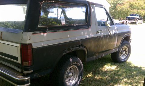 1985 Ford Bronco - A two-tone Ford Bronco is parked on grass, with another truck visible in the background.