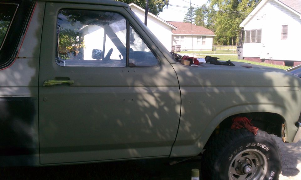 Olive green Ford Bronco side view, showing door, window, and tire. Some parts are disassembled