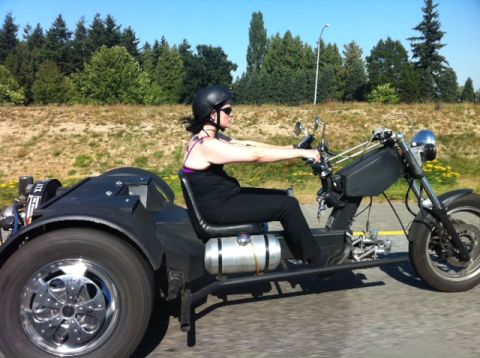 1965 Ford Fairlane - A woman rides a custom black trike motorcycle on a sunny day.