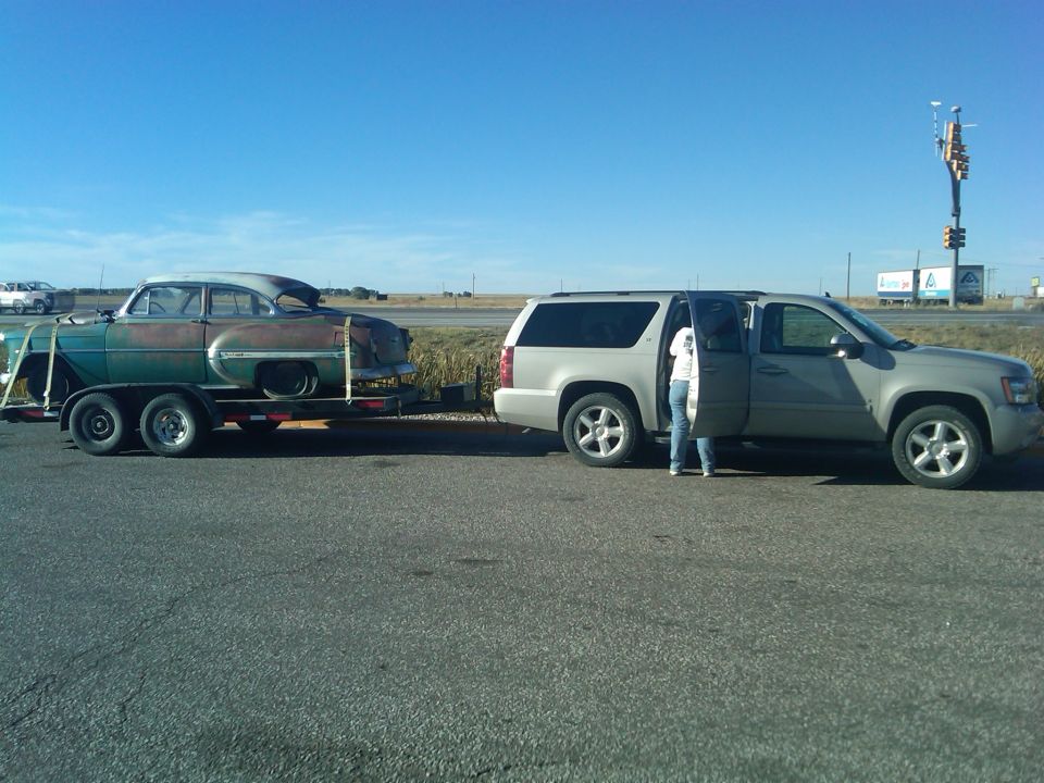 Old car on trailer, towed by silver SUV. Person standing by SUV door