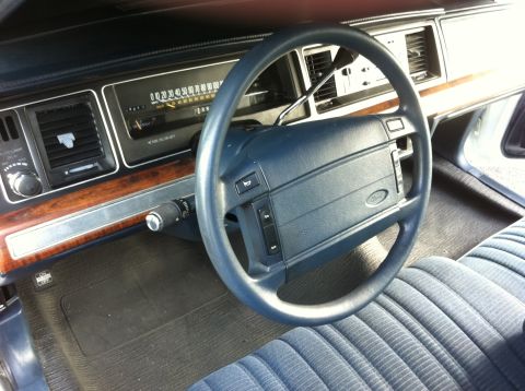 1991 Ford LTD Crown Victoria - Driver's view of a Ford sedan interior with blue cloth seats, woodgrain dash trim, horizontal speedometer and a Ford-logo steering wheel.