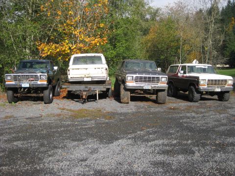 1979 Ford Bronco - Four Ford Bronco and F-Series trucks are parked in a gravel lot with trees in the background.