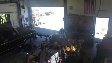 1979 Ford Bronco - A black 1978 Ford Bronco is elevated on jack stands inside a cluttered garage, with a forklift visible outside the open bay door.