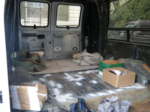 1988 Ford E350 - The rear interior of a van shows a cargo area with blankets and boxes, and some stickers on the doors.