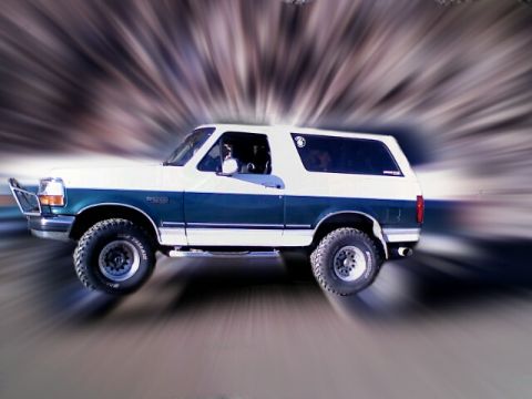 1996 Ford Bronco - A two-tone white and teal Ford Bronco SUV is captured in motion, with a visible fender badge indicating its model year.