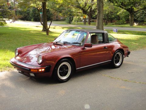 1986 Porsche 911 - A maroon 1986 Porsche 911 Targa is parked on a paved surface with trees and a street in the background.