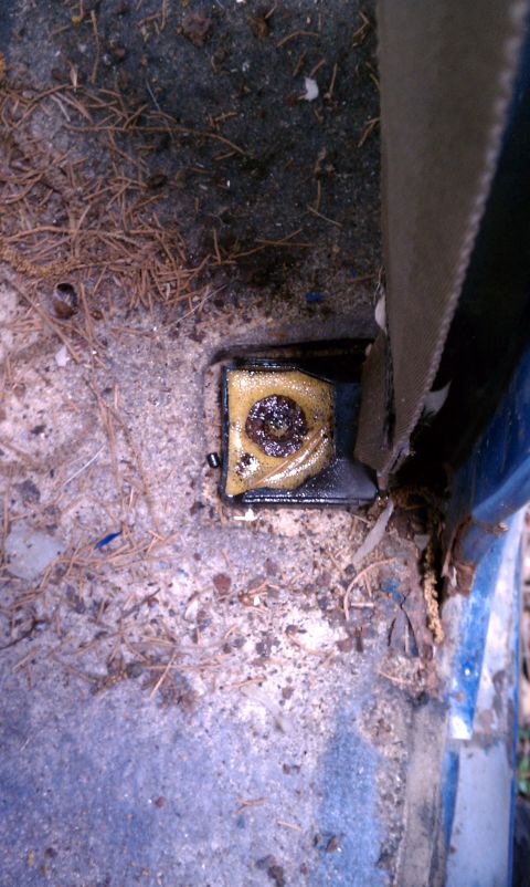 1979 Ford Bronco - Close-up of a corroded seatbelt anchor on a vehicle's floor, showing debris and a website logo.