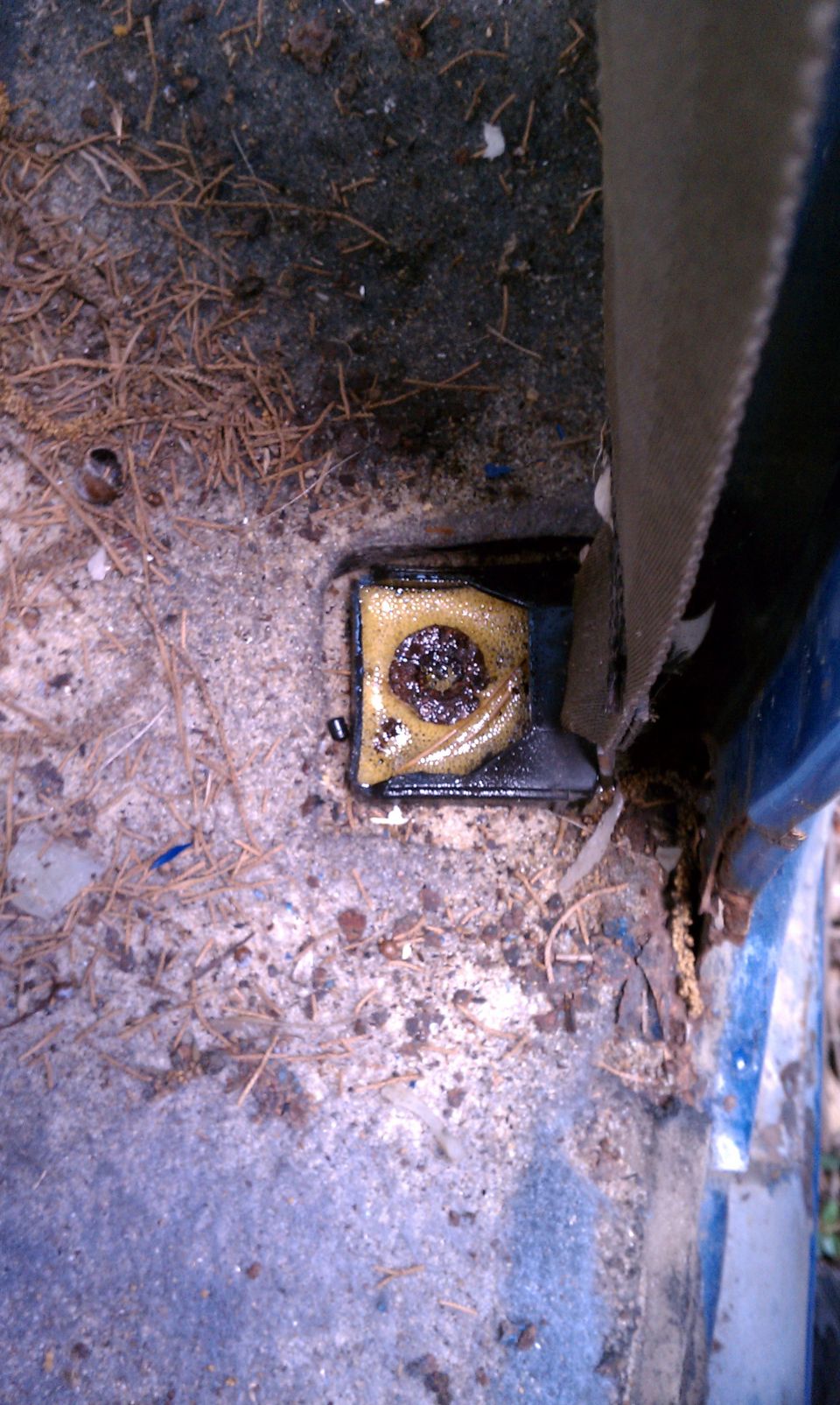 Seatbelt anchor point on a dirty vehicle floor with debris and a visible SuperMotors logo