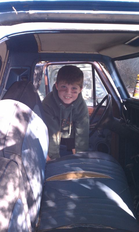 1979 Ford Bronco - A young boy with dirt on his face smiles from the interior of a blue vehicle, revealing worn and torn seats.