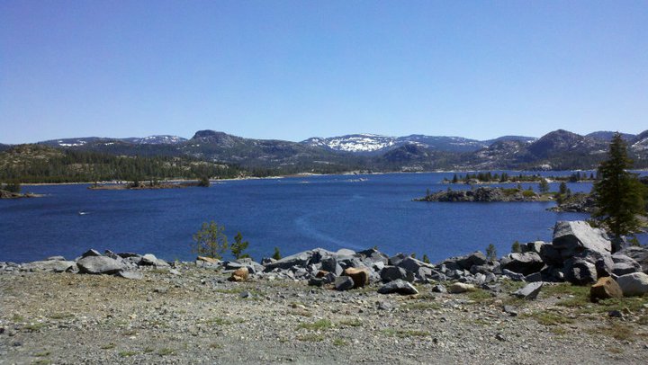 Mountain lake with trees and rocky shore under a clear blue sky. Watermark reads SUPERMOTORS