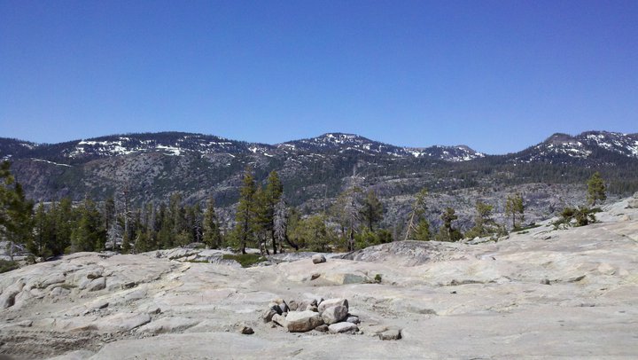 Rocky terrain with scattered trees and snow-capped mountains under a clear blue sky. A small fire pit is visible in the foreground