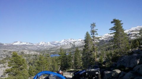 1971 Ford Bronco - Off-road vehicles are parked on a rocky incline, overlooking a lake and snow-covered mountains under a clear blue sky.