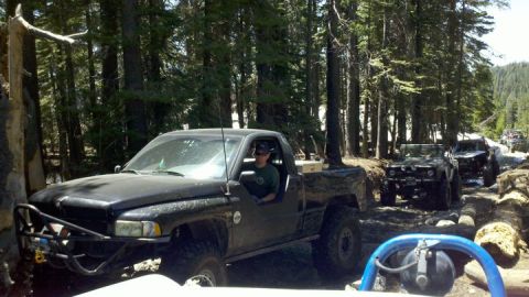1971 Ford Bronco - A line of off-road vehicles navigates a rocky, wooded trail, with a black pickup truck leading the pack.
