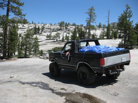 1971 Ford Bronco - A black Ford Bronco with off-road tires is parked on a rocky surface, its bed covered with a blue tarp.