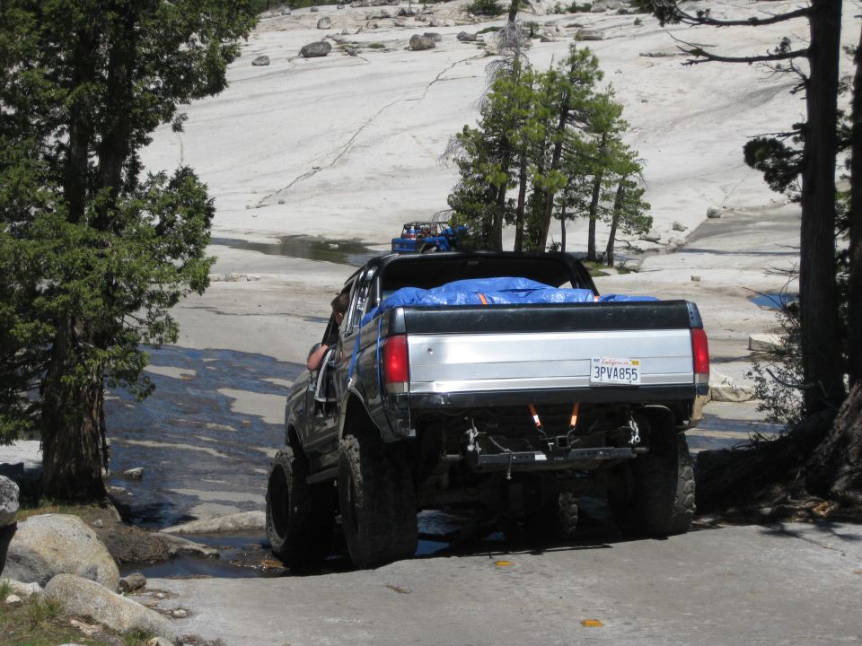 Modified pickup truck with blue tarp in bed, driving on rocky ground. Another vehicle visible in distance