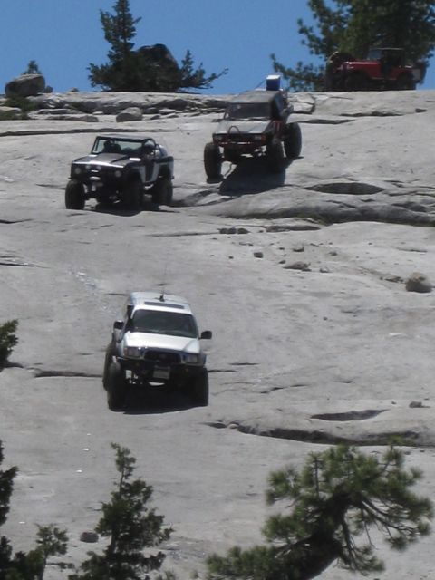 1971 Ford Bronco - Several modified off-road vehicles navigate a steep, rocky incline under a clear blue sky.