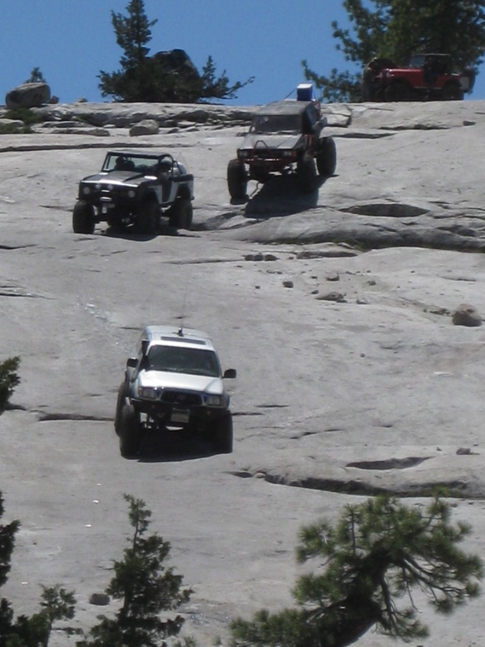 three off-road vehicles climbing a large rock face, one in front, one behind it, and one further up the slope