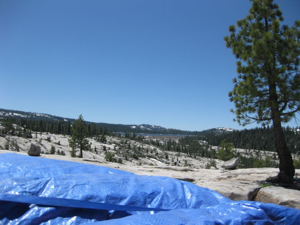 Blue tarp covers foreground in a rocky, mountainous landscape with pine trees and a distant lake