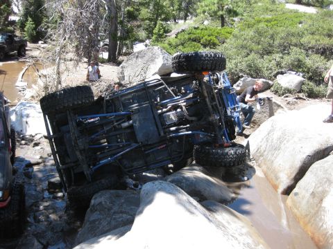 1971 Ford Bronco - An off-road vehicle has overturned in a rocky stream bed, with onlookers nearby.