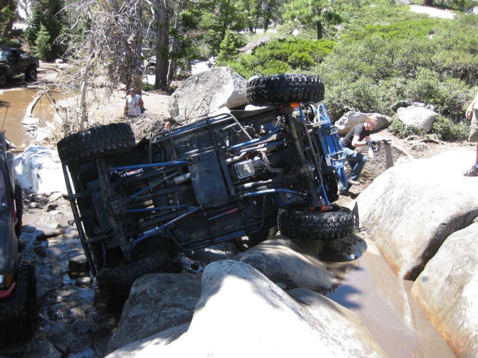 Overturned off-road vehicle on rocks in a stream, with another vehicle visible to the left