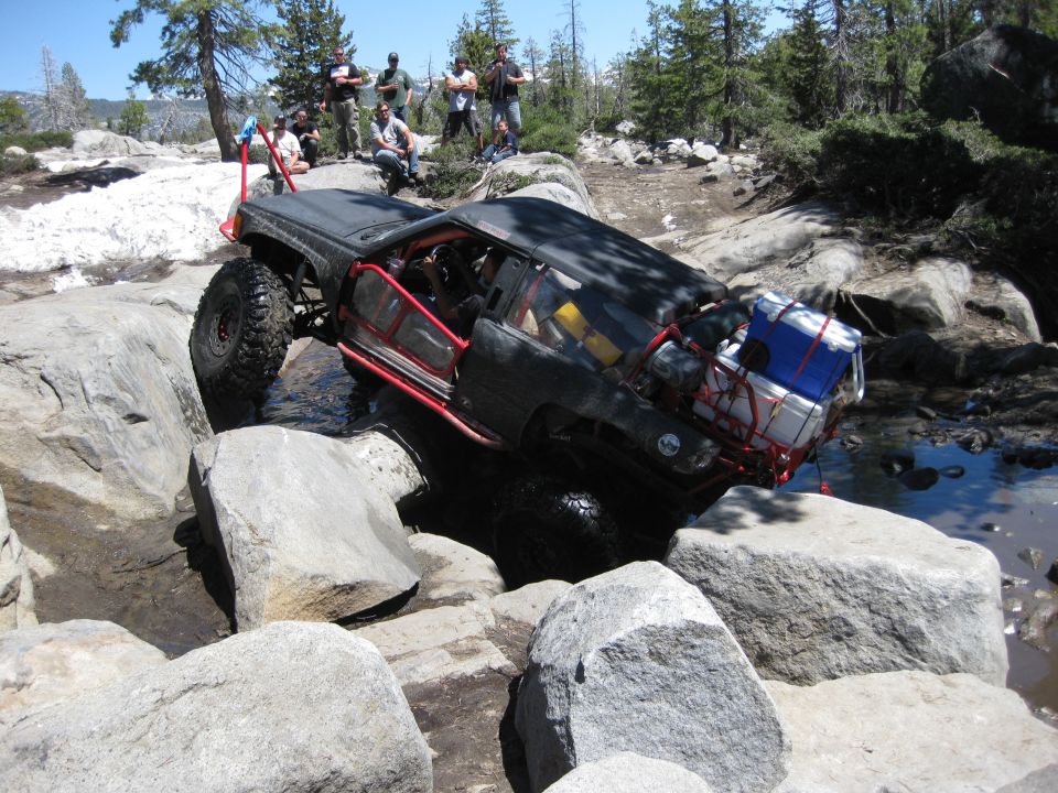 Black off-road vehicle with red roll cage navigating rocks and water