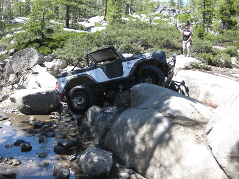 1971 Ford Bronco navigating rocky terrain with a person standing on a rock in the background