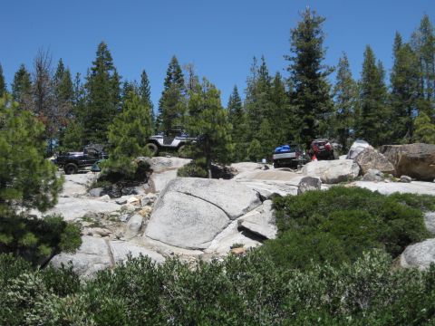 1971 Ford Bronco - Multiple off-road vehicles are parked and driving on a rocky trail surrounded by pine trees.