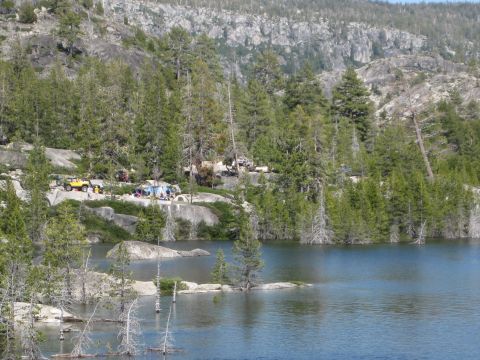 1971 Ford Bronco - A yellow Jeep and tents are set up at a campsite next to a calm lake with trees and rocky hills in the background.