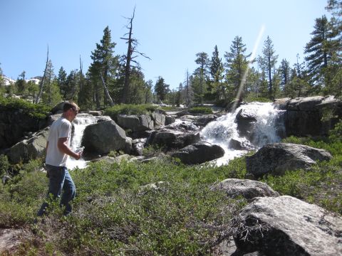 1971 Ford Bronco - A man walks near a waterfall in a rocky, wooded landscape under a clear blue sky.