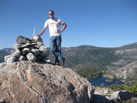 1971 Ford Bronco - A man poses on a rocky summit with a cairn, overlooking a mountain lake and forested hills under a clear blue sky.