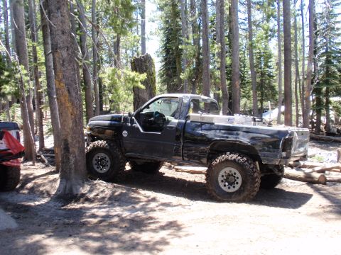 1971 Ford Bronco - A black pickup truck with large off-road tires is parked among trees, with another vehicle partially visible to the left.