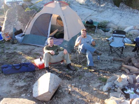 1971 Ford Bronco - Two men sit in folding chairs beside a dome tent and gear at a rocky outdoor campsite in daylight.