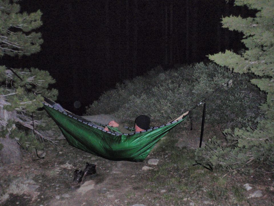 Person relaxing in a green hammock suspended between trees at night. A 'Supermotors.net' watermark is visible in the lower right corner