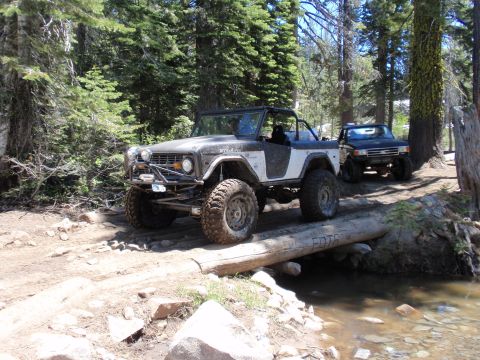 1971 Ford Bronco - Two off-road vehicles navigate a wooded trail, crossing a log over a shallow stream.