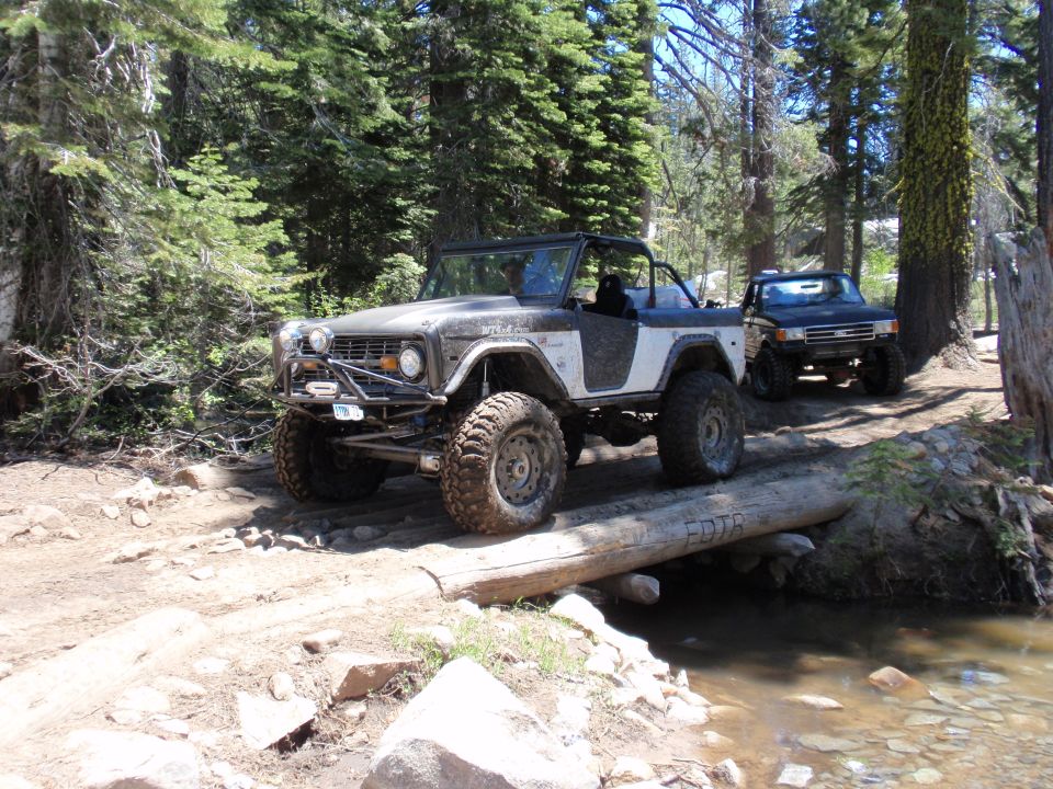 Gray and white Ford Bronco with large tires crossing a log bridge over a stream, with a black pickup truck behind it