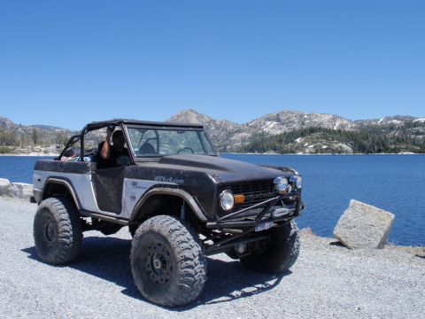 1971 Ford Bronco - Modified 1971 Ford Bronco with large off-road tires parked on a gravel shore next to a blue lake.