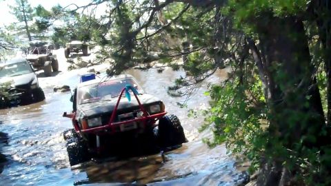 1971 Ford Bronco - Multiple off-road vehicles navigate a rocky stream bed surrounded by trees.