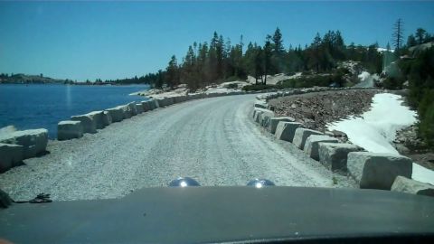 1971 Ford Bronco - View from inside a vehicle driving on a gravel road alongside a large lake with snow patches on the right.