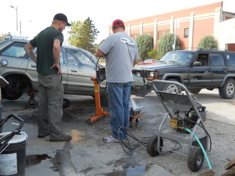 2006 Ford E350 - Men cleaning a car engine with a pressure washer, with a Jeep Cherokee in the background.
