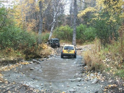 1979 Ford Bronco - Two SUVs navigate a water crossing on a wooded trail during the fall season.
