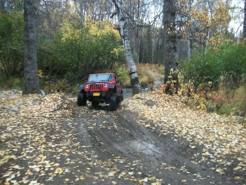 Red Jeep Wrangler driving on a muddy, leaf-covered trail in a wooded area